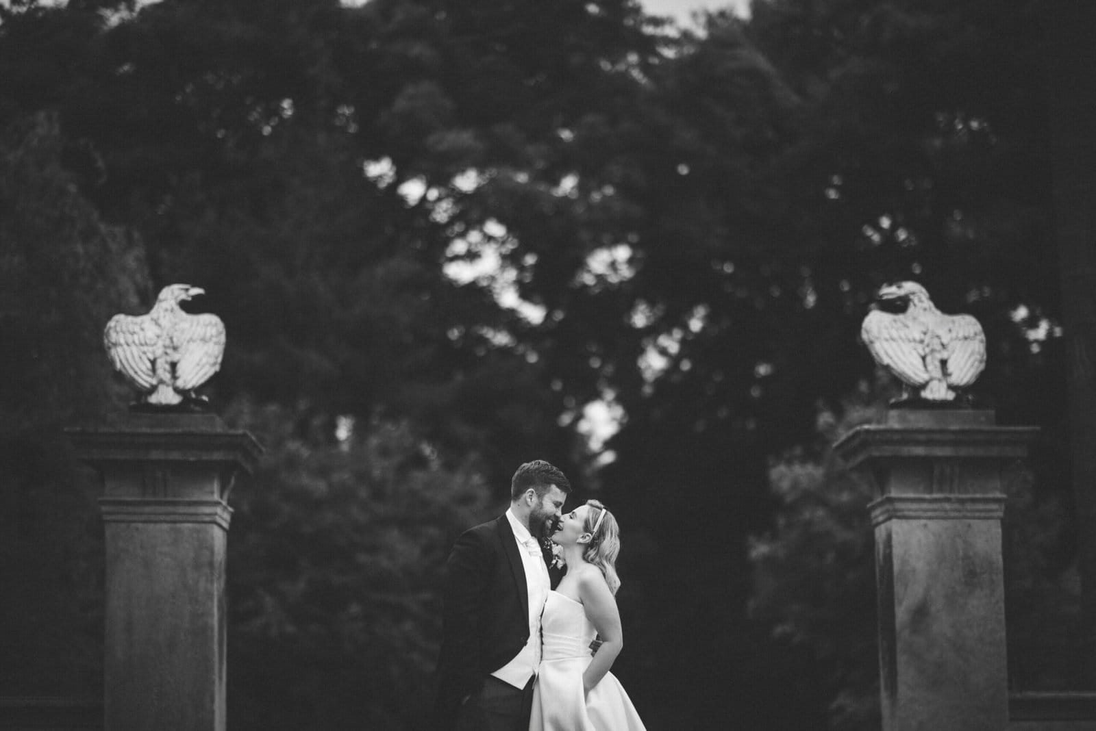 A bride and groom stand facing each other outdoors, surrounded by trees. Two stone pillars topped with eagle statues frame the couple. Captured in black and white by a Bagden Hall wedding photographer, the scene feels romantic and timeless.