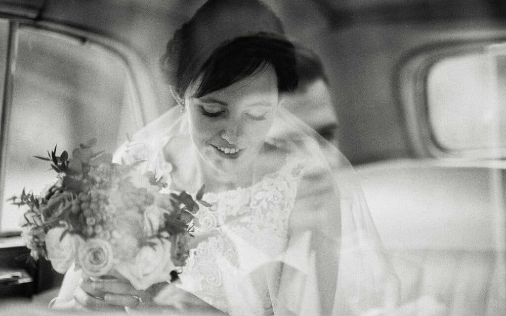 A black and white double exposure photo of a bride holding a bouquet and smiling, with a faint overlay of another person beside her in the backseat of a car.