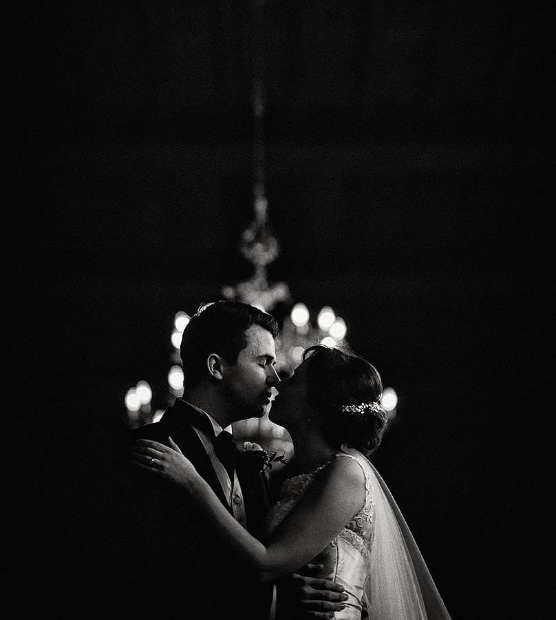 A bride and groom share an intimate moment, facing each other closely in a dimly lit setting, with a soft chandelier glowing in the background.