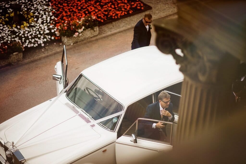 A man in a suit steps out of a white vintage car, adjusting his bow tie, while another man in a suit stands nearby. The scene is outdoors near a flowerbed and a decorative pillar.
