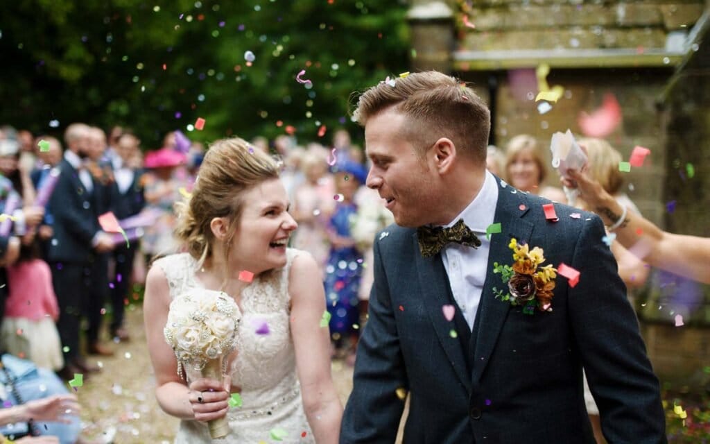 A bride and groom smile joyfully as colorful confetti is thrown over them by guests during an outdoor wedding celebration. The bride holds a bouquet and they walk hand in hand, surrounded by cheering friends and family.