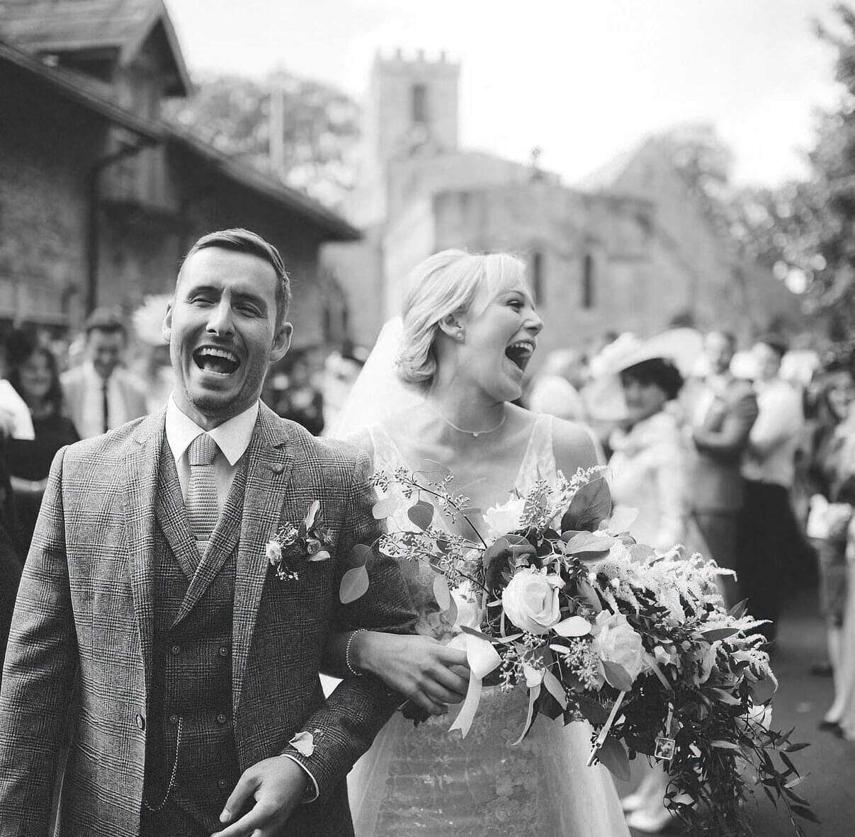 A black and white photo of a joyful couple walking arm in arm, surrounded by smiling guests at a Yorkshire wedding. The bride holds a large bouquet of flowers, and both are laughing. In the background, capture the charm with an old stone building and trees, perfect for any wedding barn photographer.