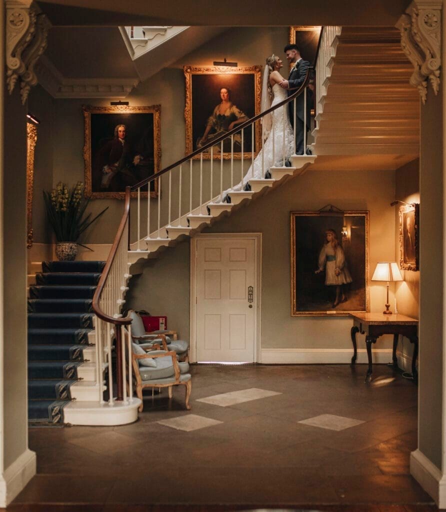 A couple embraces on a grand staircase in an elegant room with classic portraits. The Norwood Park wedding photographer captures the moment as the dark carpet and vintage furniture—including two chairs and a lamp—enhance the warm, historic ambiance.