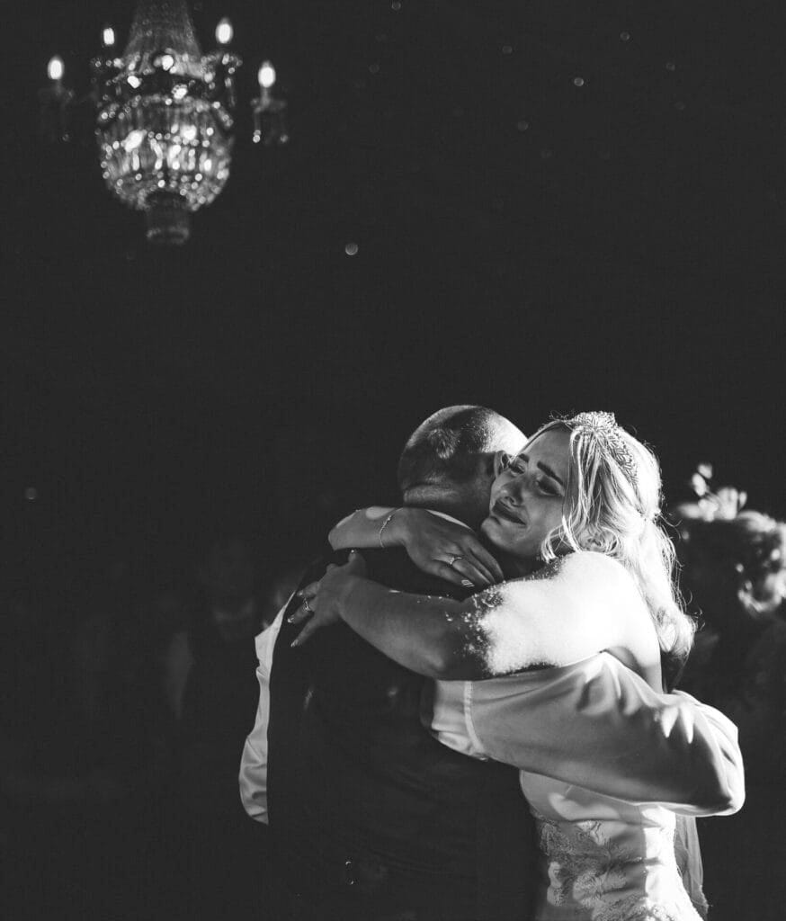A bride in a wedding dress and a man in a suit embrace closely while dancing, captured beautifully by Norwood Parks wedding photographer. Soft lighting highlights their moment beneath a chandelier, enhancing the intimate and emotional atmosphere against the dark, out-of-focus background.