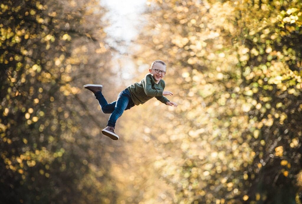 A young boy wearing glasses, a green hoodie, and jeans is midair, smiling and appearing to be flying or jumping, surrounded by sunlit, blurred autumn trees in the background.