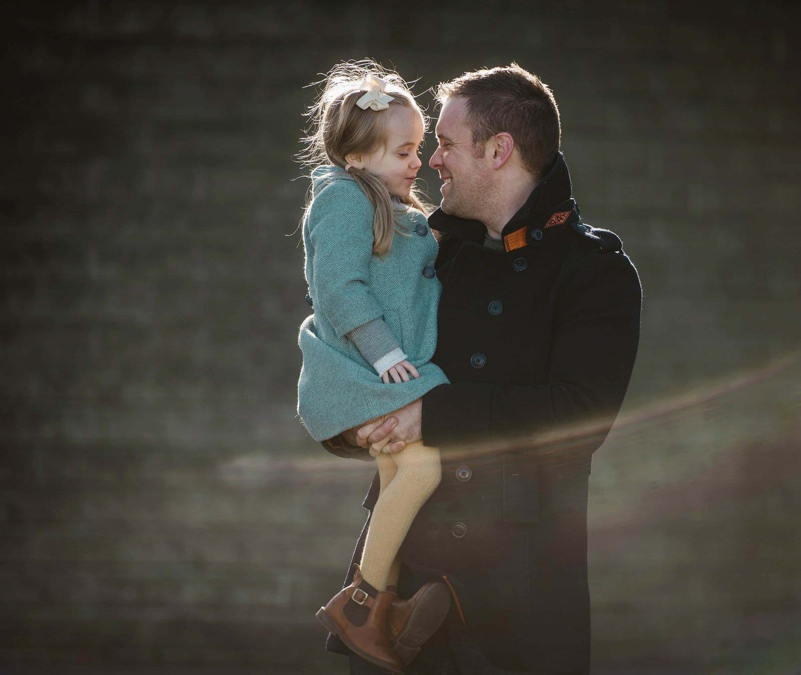 A man in a dark coat holds a smiling young girl in a blue dress with a white bow in her hair. They look at each other affectionately with blurred greenery in the background. Sunlight creates a soft lens flare.