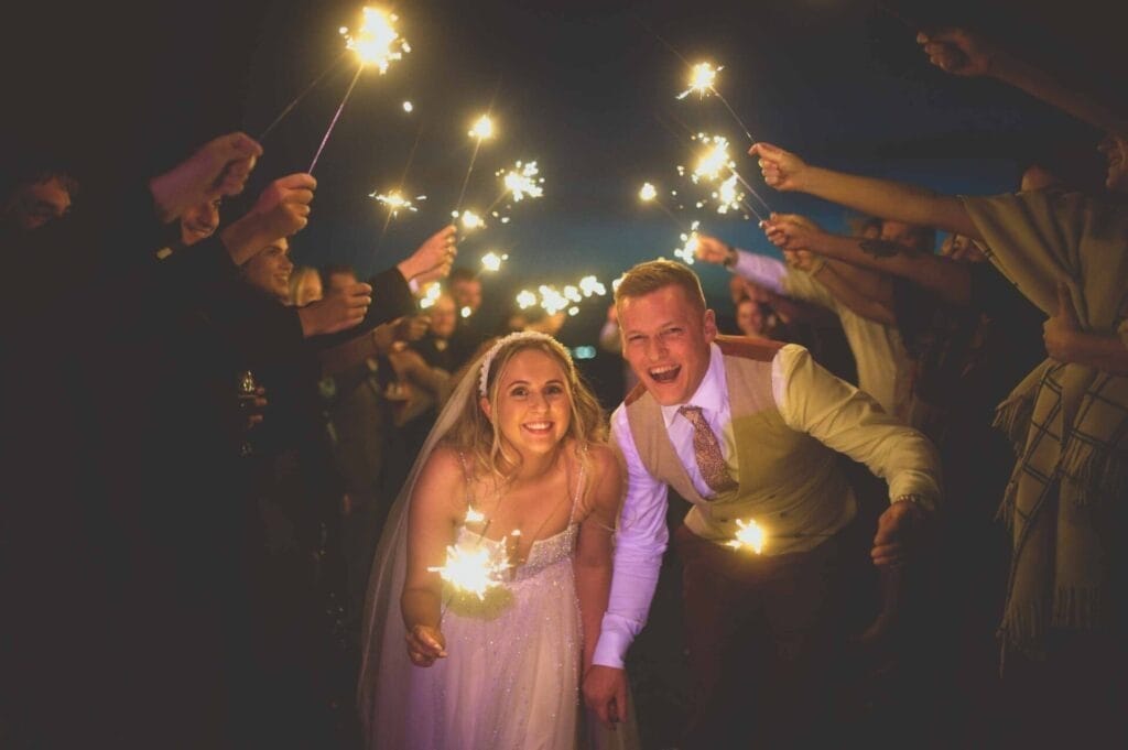 A joyful bride and groom walk through a tunnel of sparklers at their Little Wold Vineyard wedding. The bride, in her white dress and veil, and the groom, in his light-colored vest and tie, smile widely amidst the festive atmosphere captured in this enchanting photo.