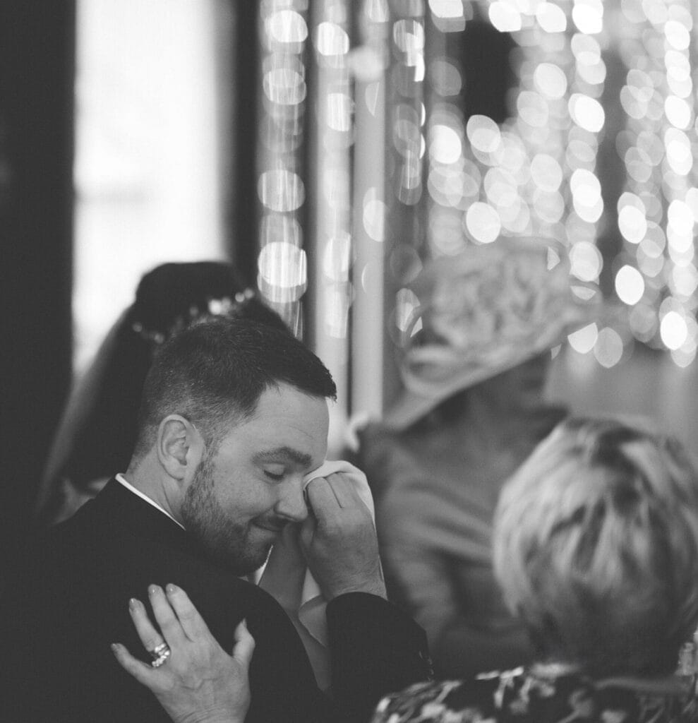 A black-and-white photo captures a poignant moment at Hornington Manor. A man in a suit wipes tears with a tissue, surrounded by elegantly dressed guests. Bokeh lights twinkle in the backdrop, as a comforting hand rests on his shoulder—a timeless Hornington Manor wedding photo captured forever.