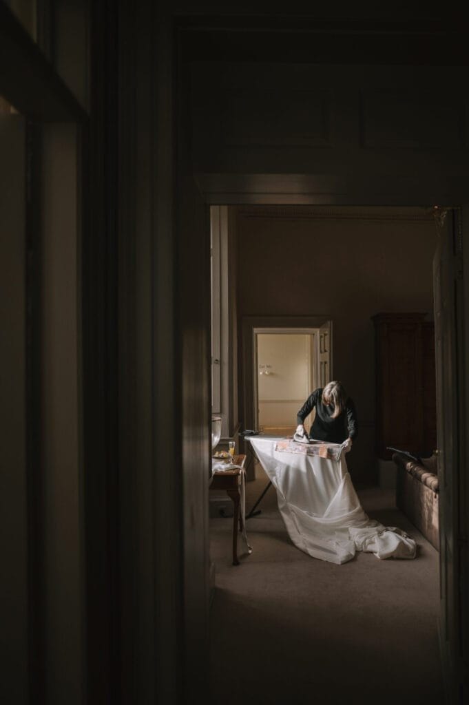 A person stands at a table, working with white fabric and sewing tools in a dimly lit room, with soft light coming through a tall window beside them—capturing the quiet artistry of a Howsham Hall Wedding Photographer.