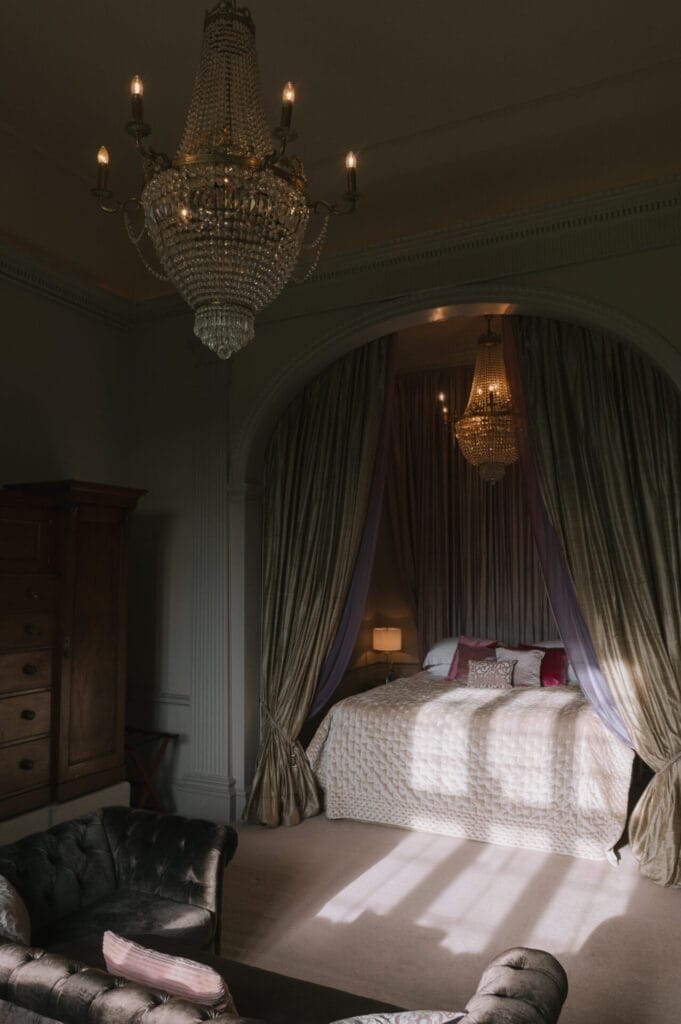 Elegant bedroom with a large bed under an arched canopy of draped curtains, two crystal chandeliers, a dark leather sofa, and sunlight streaming onto the carpeted floor—perfect inspiration for any Howsham Hall Wedding Photographer.