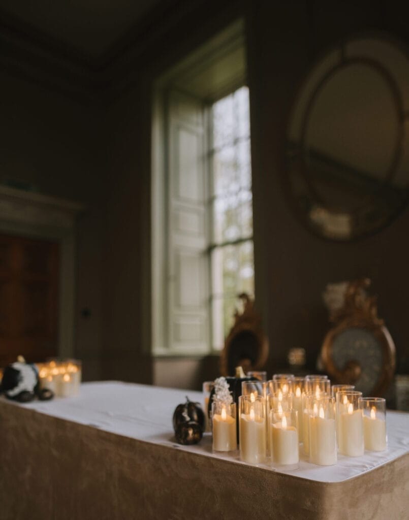 A table covered with a white cloth holds several lit candles in glass holders. The room is dimly lit, with a large window and mirrors visible in the background—perfect for a Howsham Hall Wedding Photographer to capture the calm, peaceful atmosphere.