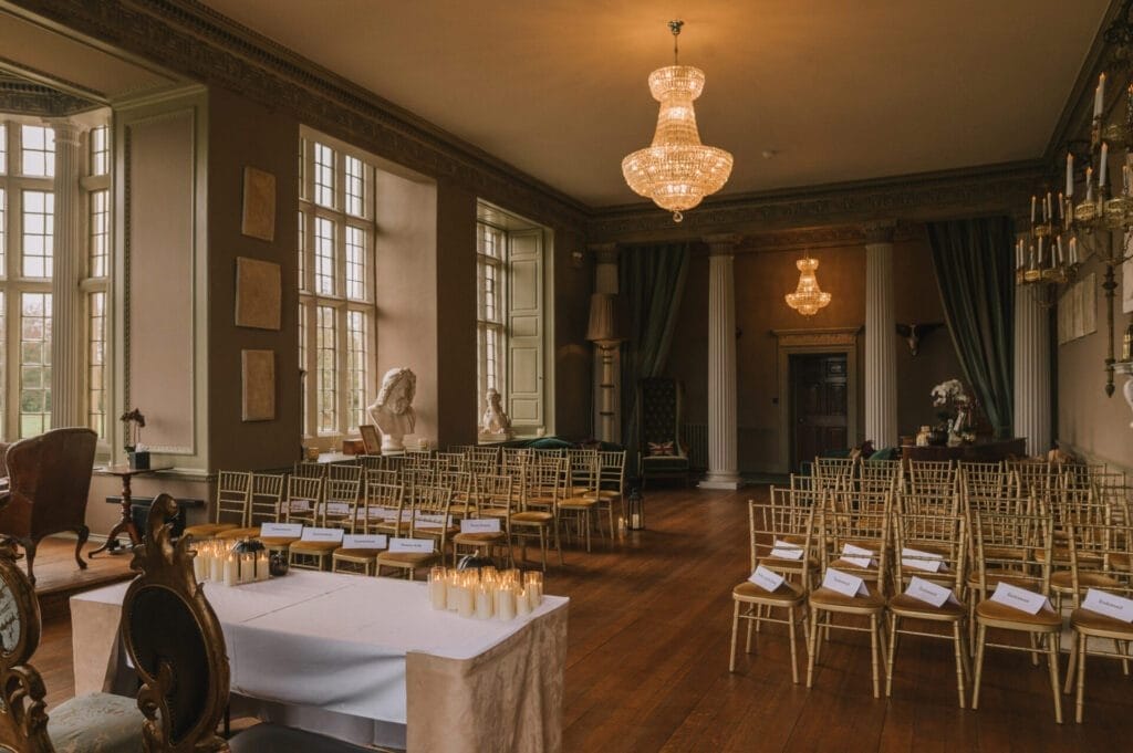 A grand, elegant room with large windows, chandeliers, gold chairs in rows, draped curtains, classical sculptures, and a table with candles—perfectly prepared for a formal event or a Howsham Hall Wedding Photographer to capture unforgettable moments.