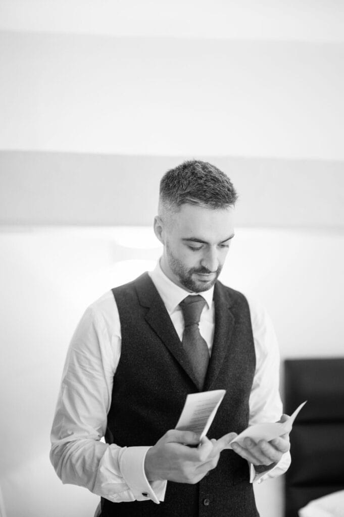 A man in a dress shirt, tie, and vest stands indoors, looking down at papers in his hands. Captured in black and white by a Howsham Hall Wedding Photographer, the background is softly out of focus.