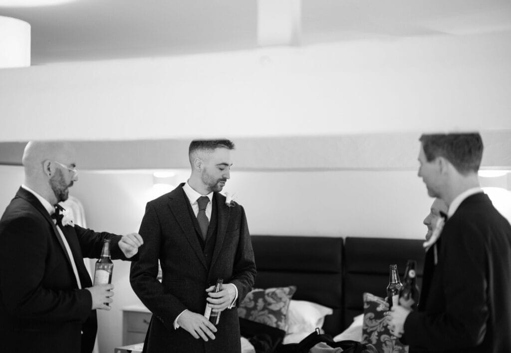 Four men in suits, holding bottles, stand talking in a bedroom with beds and pillows behind them. The relaxed mood suggests theyre preparing for a formal event—perfectly captured by a Howsham Hall Wedding Photographer. The image is in black and white.