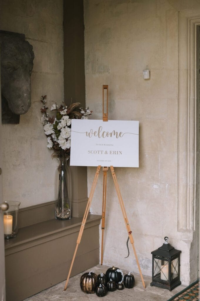 A white welcome sign on an easel reads Welcome to the wedding of Scott & Erin. Styled by their Howsham Hall Wedding Photographer, it stands near a vase with white flowers, black and gold pumpkins, candles, and a lantern against a stone wall.