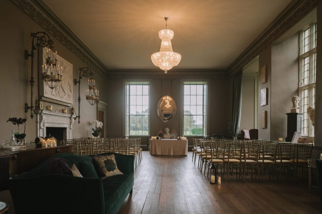 An elegant room set up for a wedding ceremony at Howsham Hall, with rows of gold chairs facing a table by large windows, a green sofa on the left, chandeliers overhead, and soft natural light—perfect for any Howsham Hall Wedding Photographer.