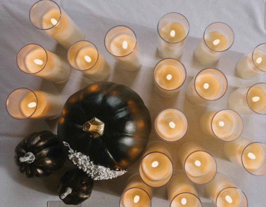 A group of lit pillar candles and two black pumpkins are arranged on a white tablecloth, creating a warm and festive display—perfect inspiration for any Howsham Hall Wedding Photographer seeking unique decor ideas.