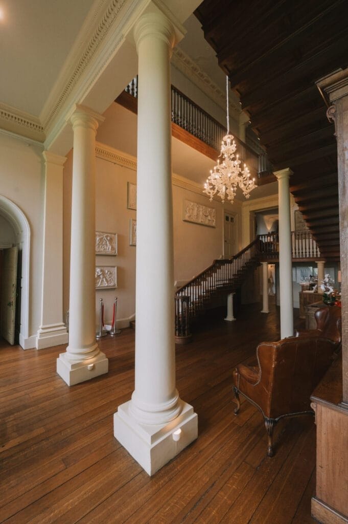 A grand interior with tall white columns, wooden floors, a dark wood staircase with a railing, a brown leather armchair, and an ornate chandelier—an inviting scene for any Howsham Hall Wedding Photographer.