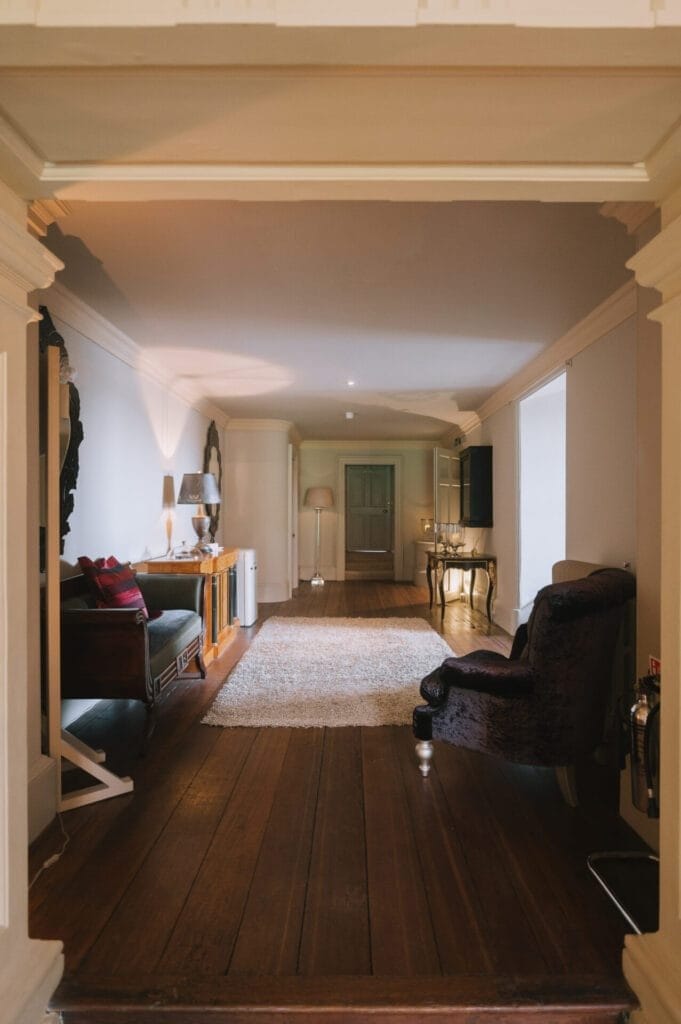 A cozy, well-lit hallway with wooden floors, a rug, an armchair, side tables, lamps, and a bench with cushions—a perfect spot for relaxed portraits by a Howsham Hall Wedding Photographer—decorated in warm, neutral tones.
