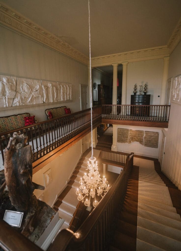 Elegant staircase with wooden railings and white steps, a crystal chandelier hanging in the center—perfect for a Howsham Hall Wedding Photographer to capture. The upper level features classical wall art, red cushions, and decorative columns.