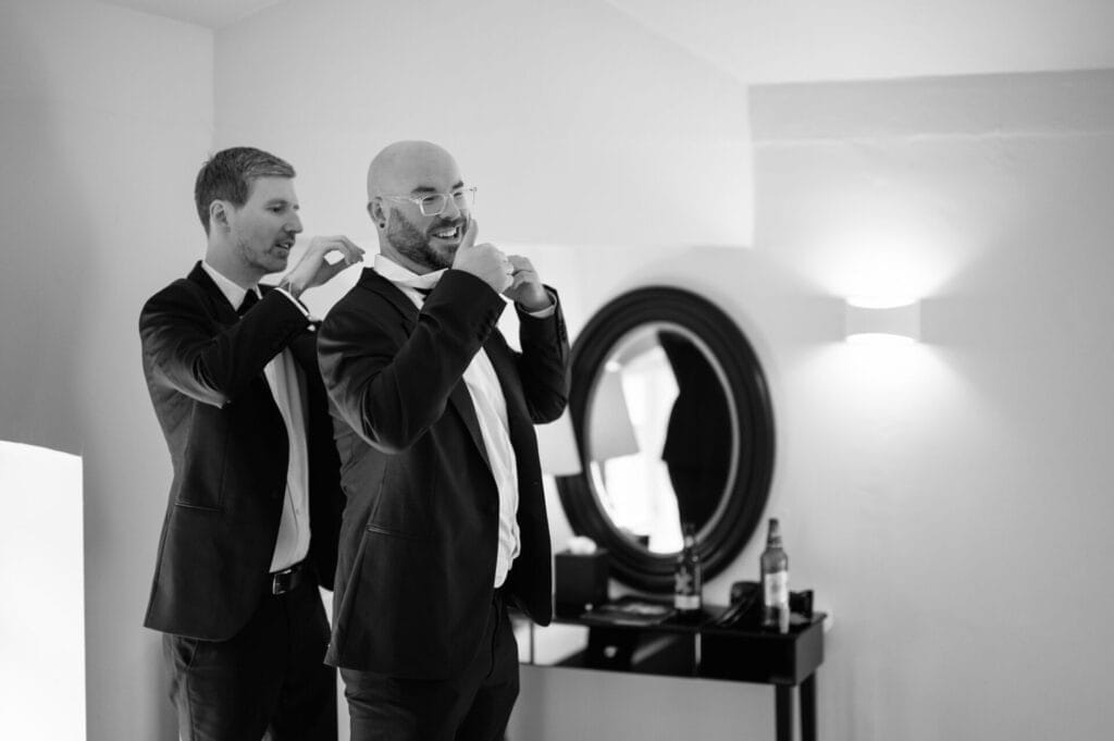 Two men in suits stand in a well-lit room; one helps the other adjust his bow tie. A round mirror, a table, and a bottle are visible. Both appear to be getting ready for a formal event—captured by a Howsham Hall Wedding Photographer.