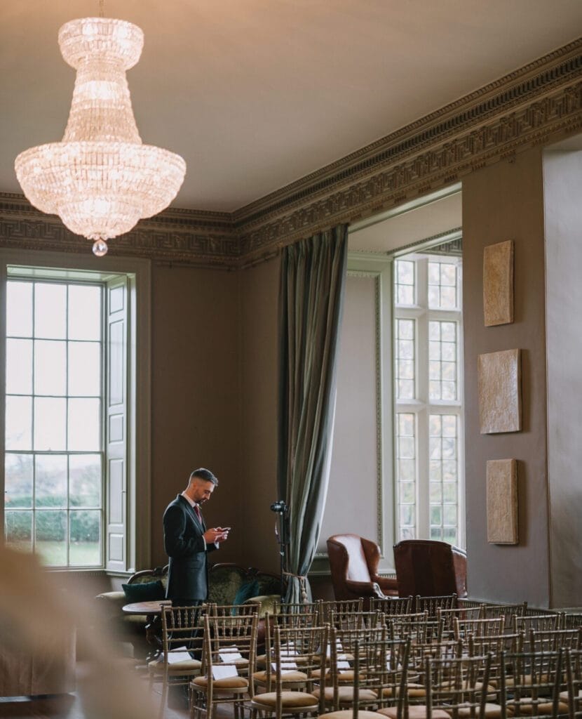 A man in a suit stands alone in an elegant room at Howsham Hall, with large windows, ornate molding, and a chandelier, looking at his phone. Rows of empty chairs are arranged in front—perfect for a Howsham Hall Wedding Photographer to capture.