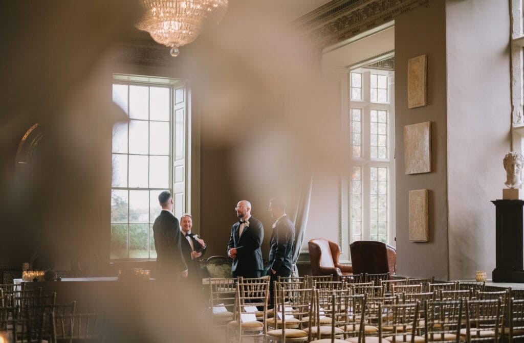 Four men in formal attire stand talking near tall windows in an elegant, softly lit room at Howsham Hall, with rows of empty gold chairs. The blurred foreground draws focus to the conversation—perfect for a Howsham Hall Wedding Photographer.