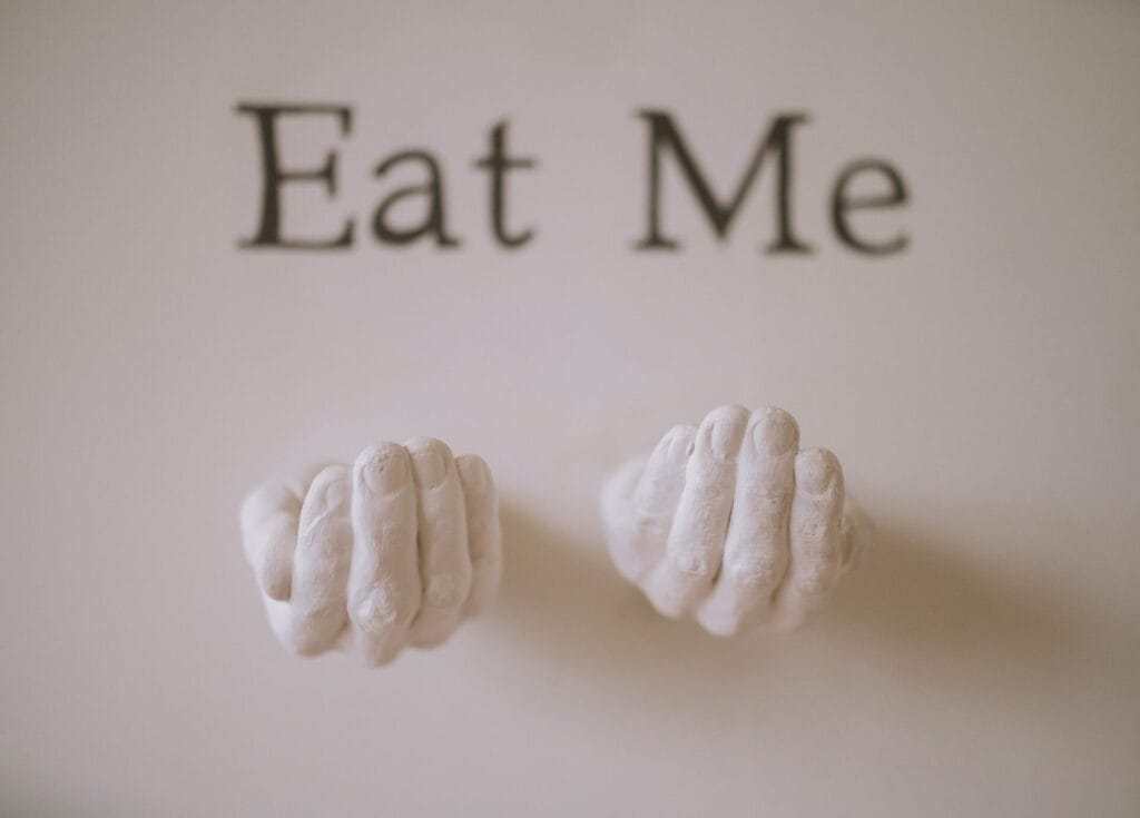 Two white sculpted hands positioned as if holding something, mounted on a wall beneath the bold black text Eat Me on a light background—a striking detail often appreciated by a Howsham Hall Wedding Photographer.