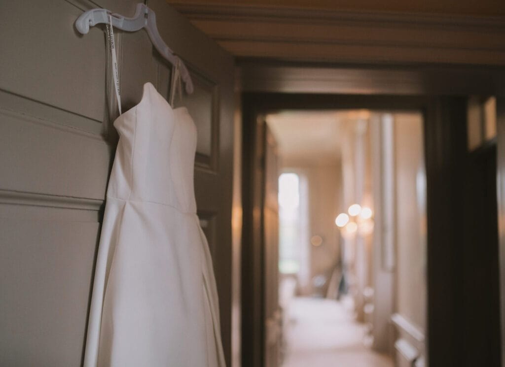 A white strapless wedding dress hangs on a door inside a softly lit, elegant hallway at Howsham Hall, with blurred lights and windows in the background—perfect for a Howsham Hall Wedding Photographer to capture.