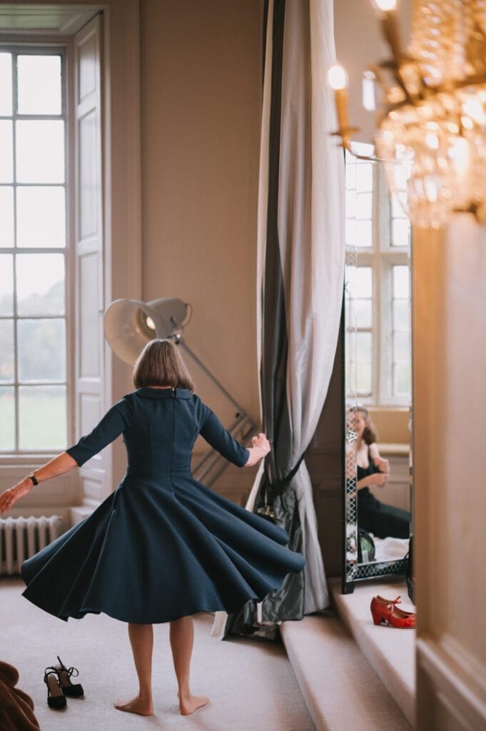 A woman in a dark blue dress twirls barefoot in a bright room with large windows. Shoes rest on the floor, her reflection partially visible in a tall mirror—an elegant moment captured by a Howsham Hall Wedding Photographer.