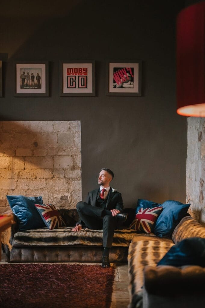 A man in a suit sits on a vintage sofa with Union Jack pillows in a cozy, dimly lit room. Art and photos by a Howsham Hall Wedding Photographer hang above him, while warm lamps softly illuminate the rustic space.