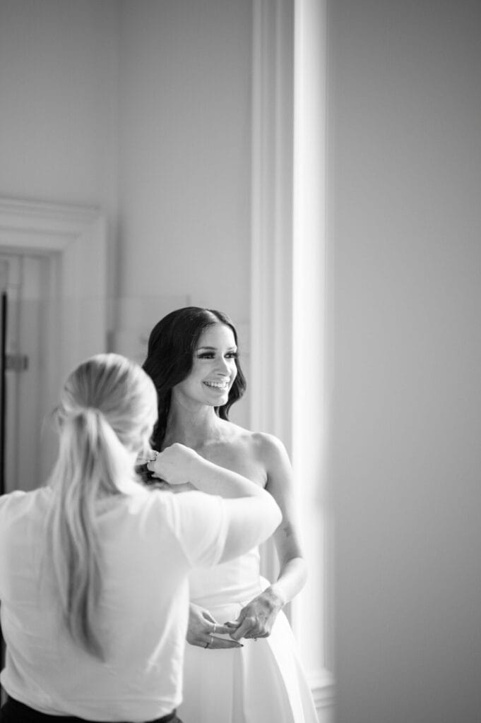 A smiling bride in a strapless dress stands near a window as another woman adjusts her hair. Captured in black and white by a Howsham Hall Wedding Photographer, the scene exudes a soft, elegant atmosphere.