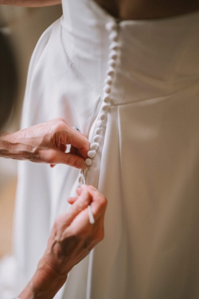 A close-up of hands fastening buttons on the back of a white wedding dress, highlighting the fabric and elegant row of fabric-covered buttons—perfect inspiration from a Howsham Hall Wedding Photographer.