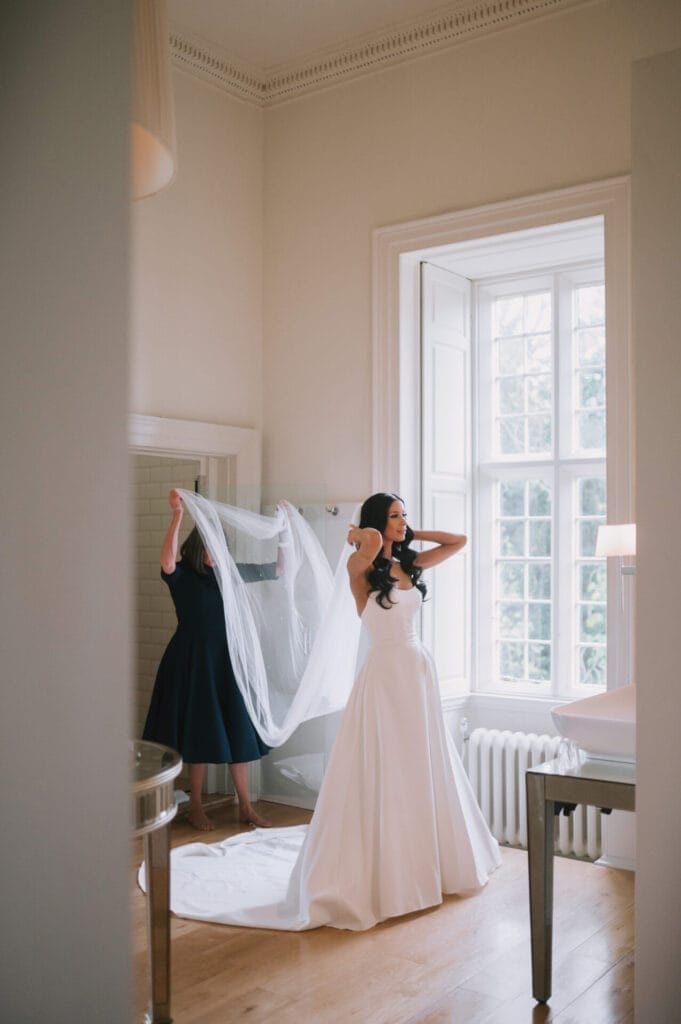 A bride in a white gown adjusts her hair while another woman helps arrange her long veil in a bright room with large windows and wooden floors, perfectly captured by a Howsham Hall Wedding Photographer.