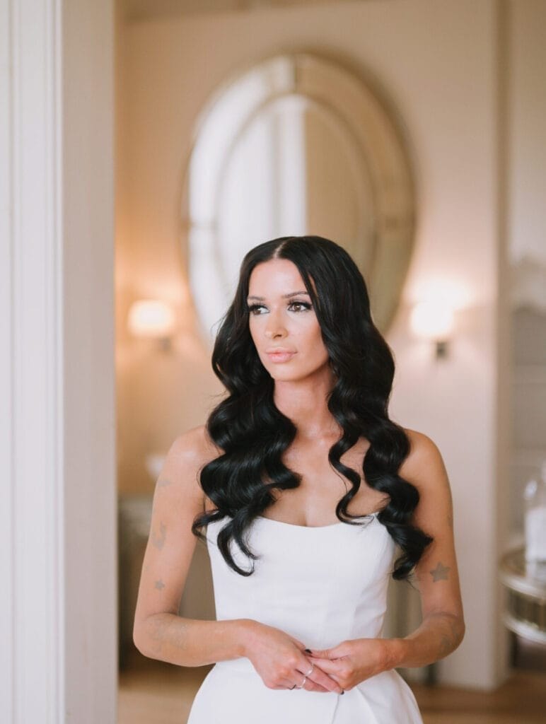 A woman with long, wavy black hair and a white strapless dress stands indoors, hands gently clasped. She looks slightly to the side with a calm expression. Soft lighting and an ornate oval mirror evoke the elegance of a Howsham Hall Wedding Photographer.