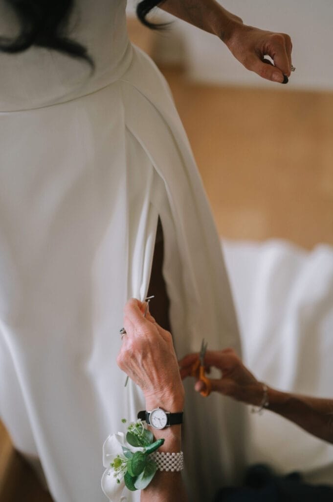 A Howsham Hall Wedding Photographer captures a person carefully cutting or adjusting the slit on a white wedding dress, while another stands nearby; the person cutting wears a watch and a corsage with greenery.
