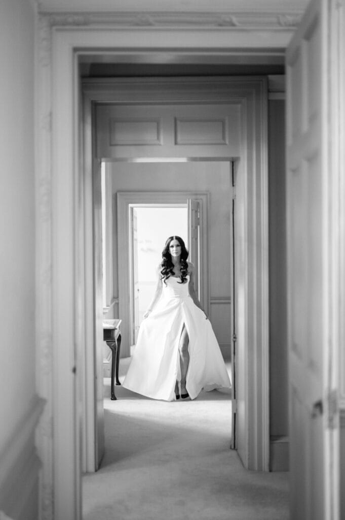 A woman in a flowing white dress stands in a doorway, framed by open doors in a softly lit, elegant interior. Captured by a Howsham Hall Wedding Photographer, the black and white image evokes a timeless and serene atmosphere.