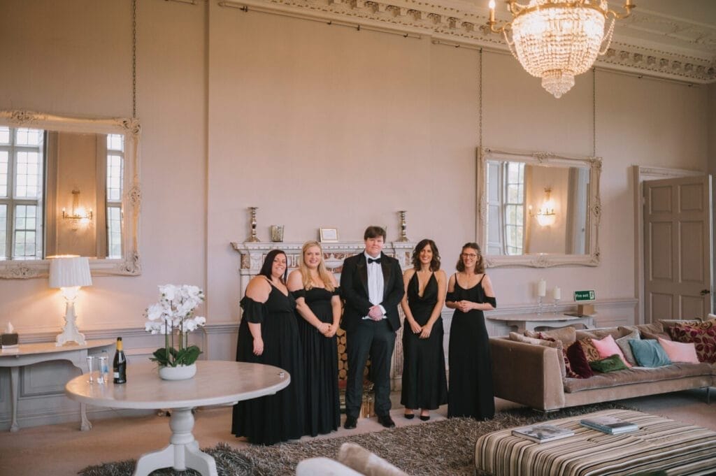 Five people dressed formally stand in a spacious, elegant living room at Howsham Hall; four women in black dresses and a man in a tuxedo pose beneath a chandelier—perfect inspiration for any Howsham Hall Wedding Photographer.
