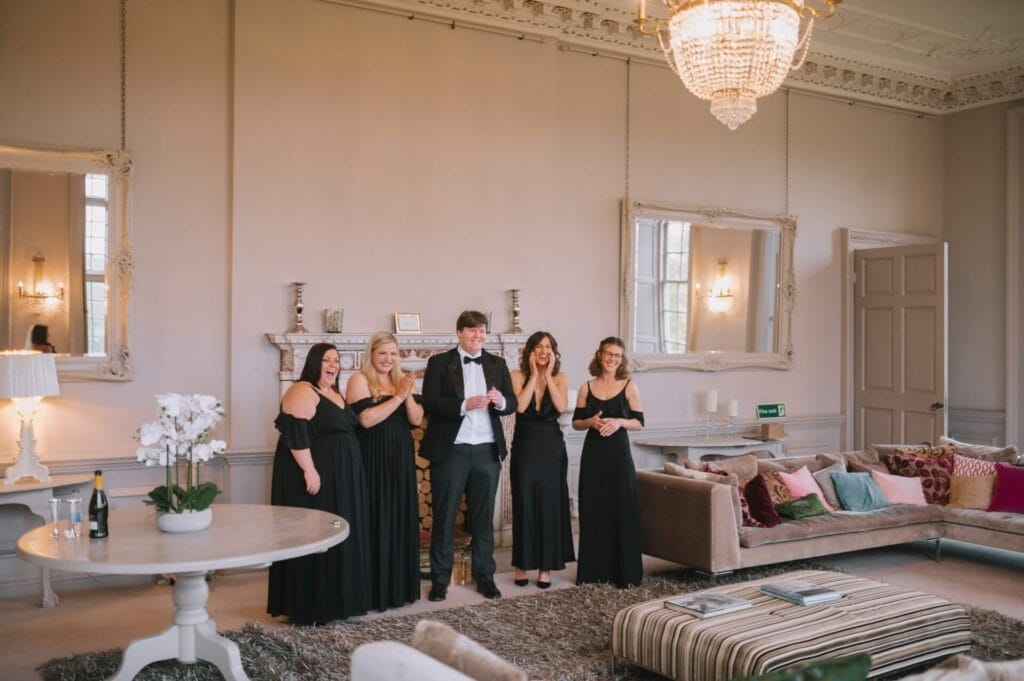 Five people, four women in black dresses and one man in a tuxedo, stand together smiling in an elegant, well-lit living room with ornate decor and chandeliers—perfect for a Howsham Hall Wedding Photographer to capture a formal celebration.