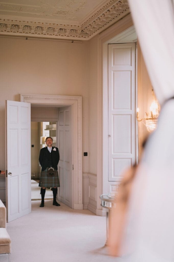 A man in a kilt stands in the doorway of an elegant, light-filled room with ornate ceilings and tall white doors, captured by a Howsham Hall Wedding Photographer as he looks towards someone out of focus in the foreground.