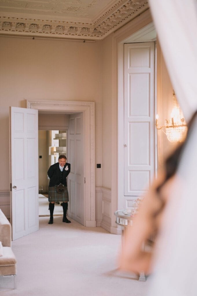 A man in a kilt stands in a doorway, smiling and wiping a tear from his eye as he sees the bride—a touching moment expertly captured by this Howsham Hall Wedding Photographer in an elegant, cream-colored room.