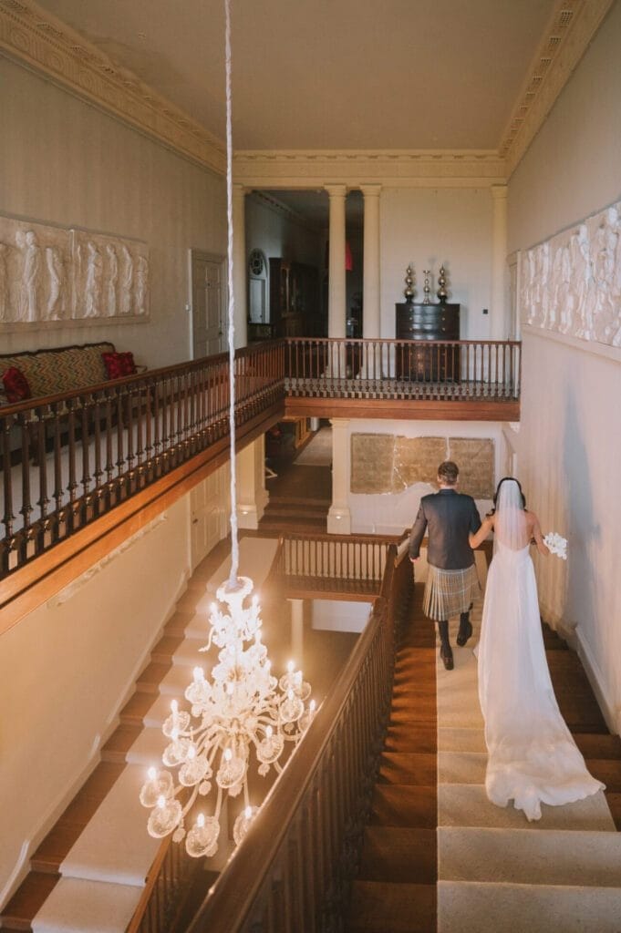 A bride in a long white dress and veil holds a bouquet and walks downstairs with a man in a kilt inside elegant, historic Howsham Hall. The scene is beautifully captured by a skilled Howsham Hall Wedding Photographer amidst ornate decor and a grand chandelier.