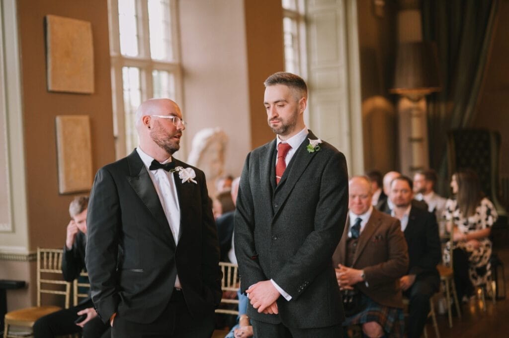 Two men in formal attire stand at the front of a room, facing each other, with seated guests in the background. Captured by a Howsham Hall Wedding Photographer, this moment exudes elegance at what appears to be a wedding or formal event.