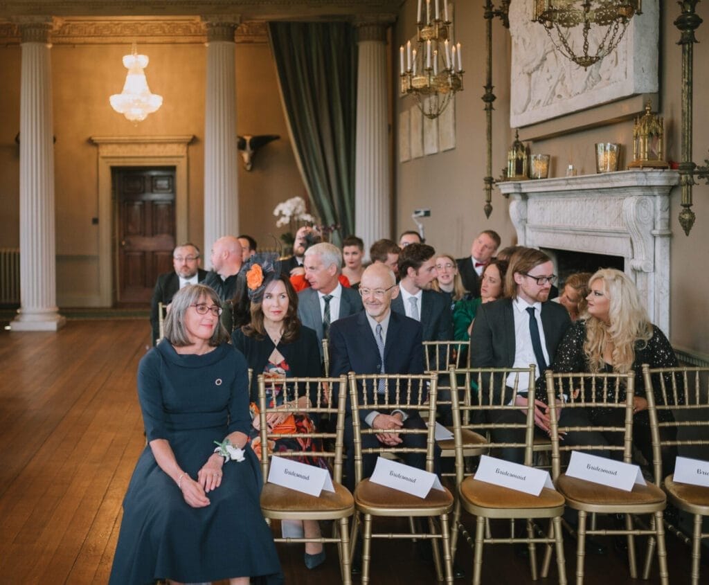 A group of people sits in elegant chairs in a formal room with chandeliers and pillars at Howsham Hall, awaiting an event. Some seats in the front row are labeled bridesmaid, and everyone is dressed in formal attire—perfect for a Howsham Hall Wedding Photographer.