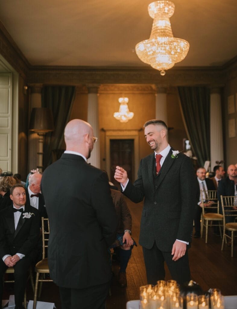Two men stand facing each other in formal attire at a wedding ceremony. One, smiling, gestures with his hand. Guests watch from rows below sparkling chandeliers in an elegant, warmly lit room—captured by a Howsham Hall Wedding Photographer.