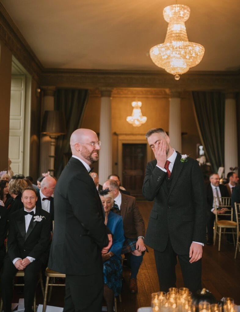 Two men in suits stand facing each other at a wedding ceremony. One smiles while the other covers his face, appearing emotional. Guests sit behind them in a grand room with chandeliers and columns, captured by a Howsham Hall Wedding Photographer.