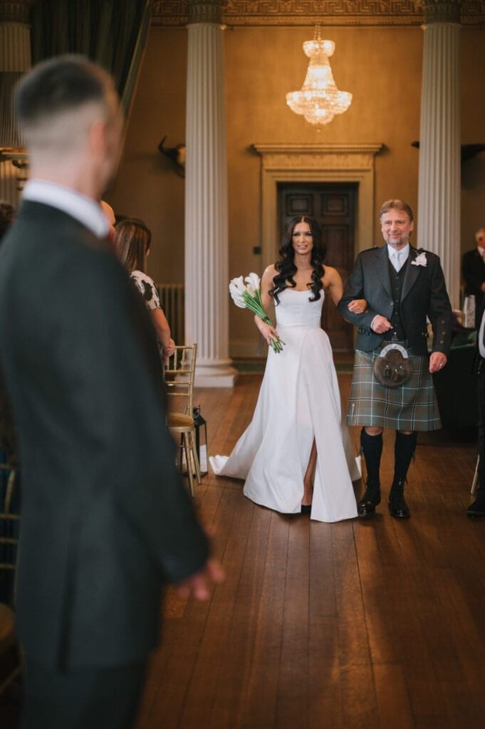 A bride in a white dress walks down the aisle holding a bouquet, accompanied by a man in a kilt, as the groom looks on in an elegant room with tall columns and a chandelier—captured by a Howsham Hall Wedding Photographer.