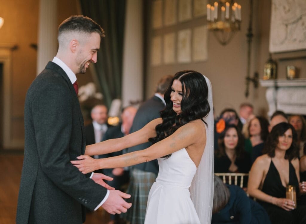 A bride in a white dress and veil smiles joyfully as she holds her groom’s hands. Captured by a Howsham Hall Wedding Photographer, they stand facing each other in front of guests at an elegant indoor ceremony.