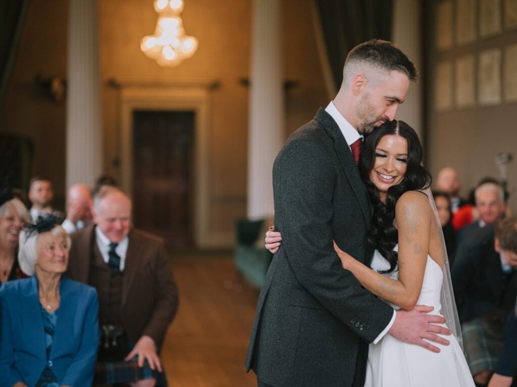 A bride and groom embrace and smile during their Howsham Hall wedding ceremony in an elegant room, with seated guests watching and smiling in the background—captured beautifully by a professional Howsham Hall Wedding Photographer.