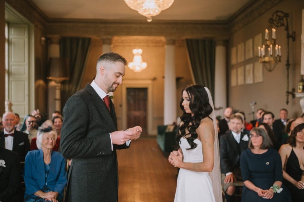A groom reads his vows to the bride during a wedding ceremony in an elegant room at Howsham Hall, surrounded by seated guests attentively watching. The bride stands facing him, wearing a white dress and veil—a perfect moment for any Howsham Hall Wedding Photographer.